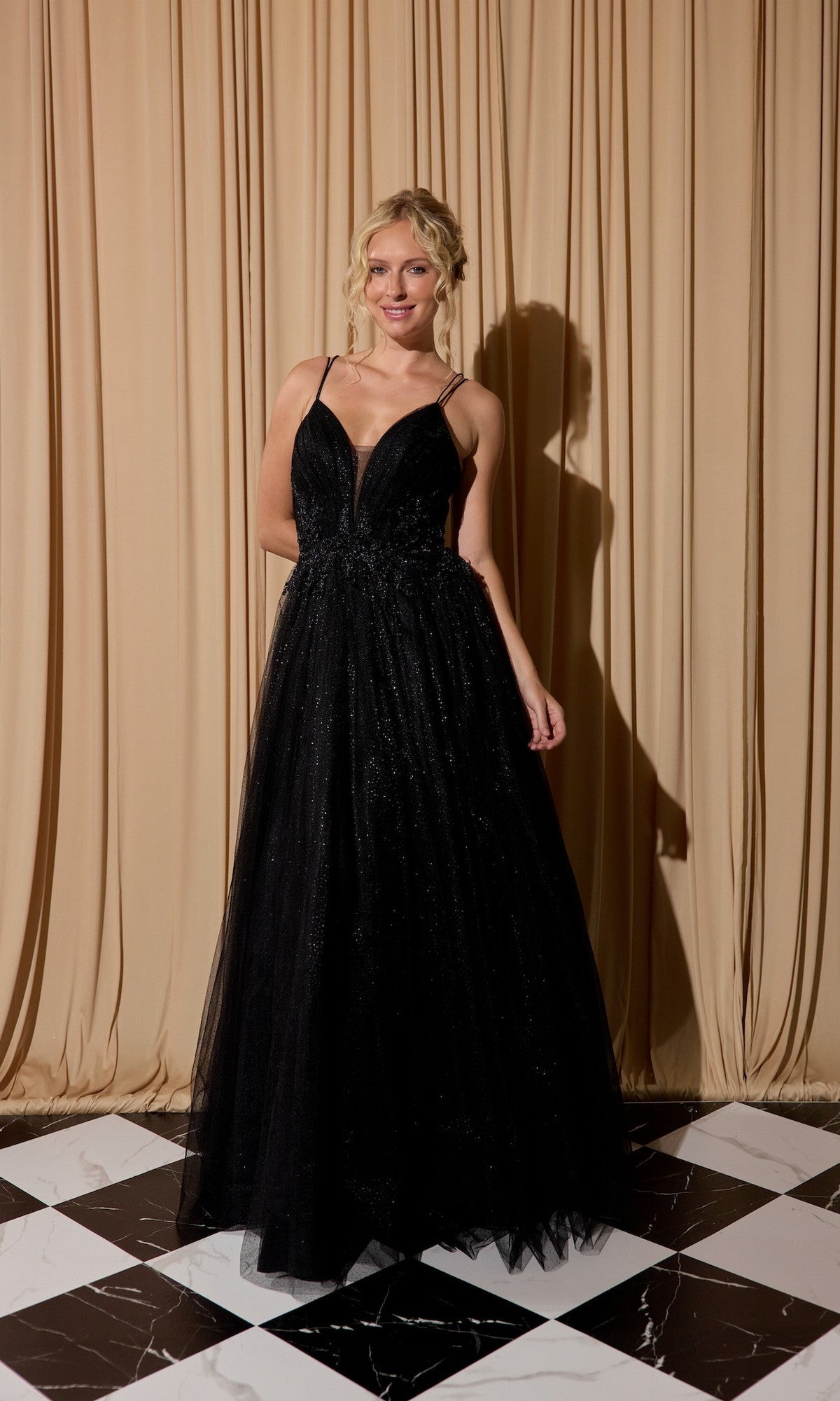 A woman wearing the Lace-Up Long Shimmer Prom Dress: Eureka 9797 stands on a black-and-white checkered floor in front of beige curtains.